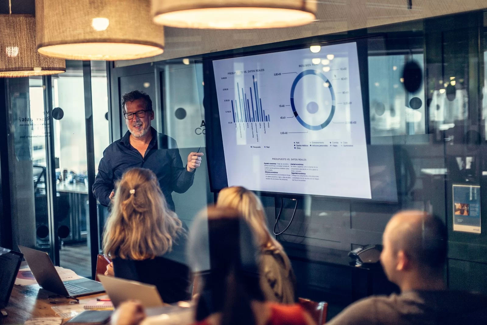 A man stands in front of a screen displaying charts and graphs, presenting to a group of people seated around a conference table in a modern office meeting room.