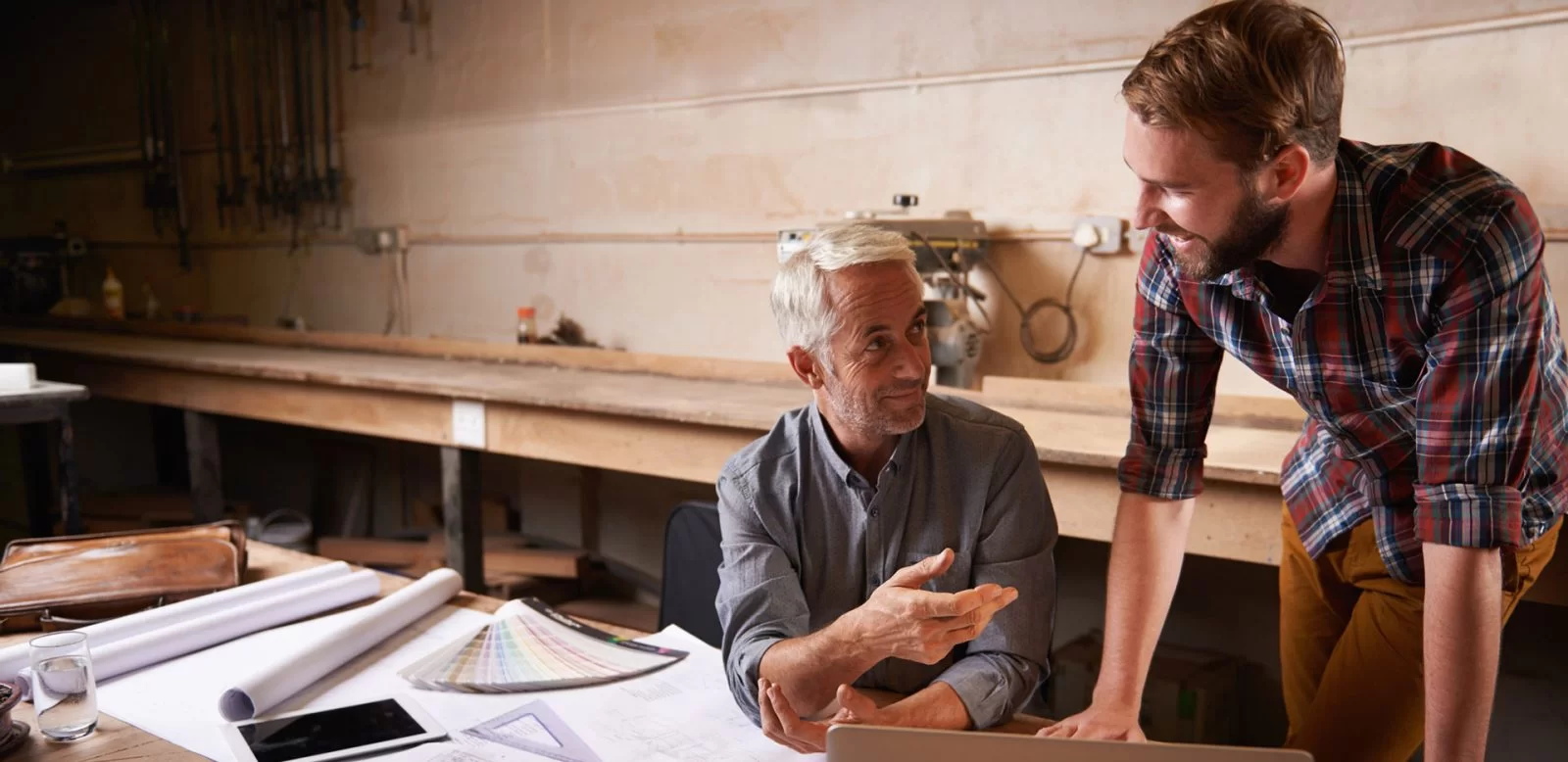 Two men working together in a workshop, one seated at a desk with papers, a laptop, and color swatches, while the other stands and smiles, engaging in conversation. Tools and equipment are visible in the background.