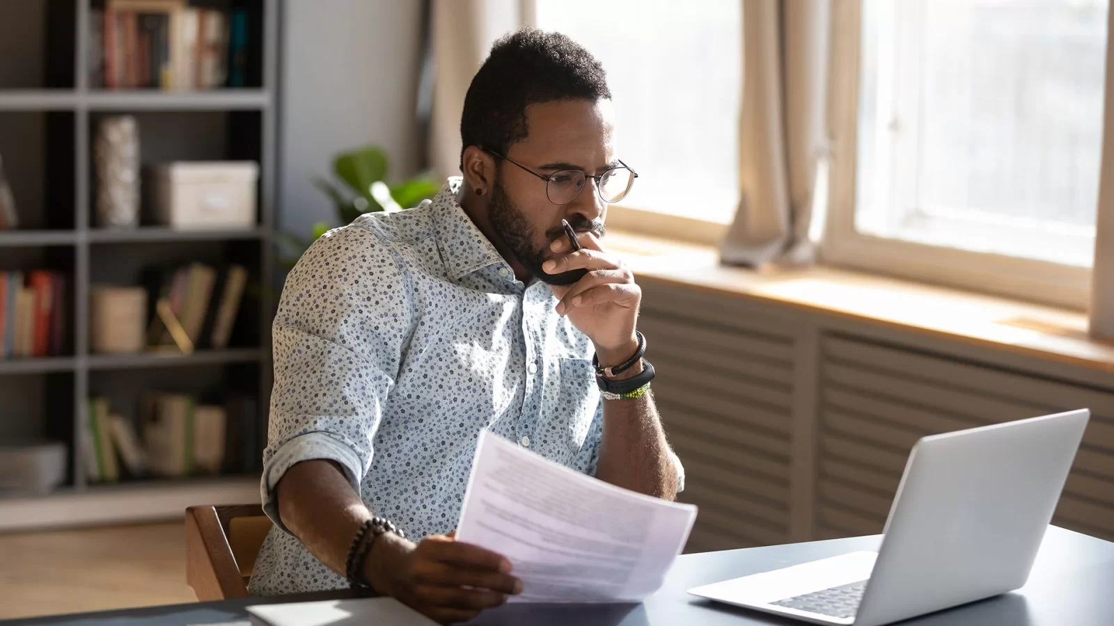 A man wearing glasses sits at a desk, looking thoughtfully at a laptop screen while holding a document in his hand. Bookshelves and large windows are visible in the background.