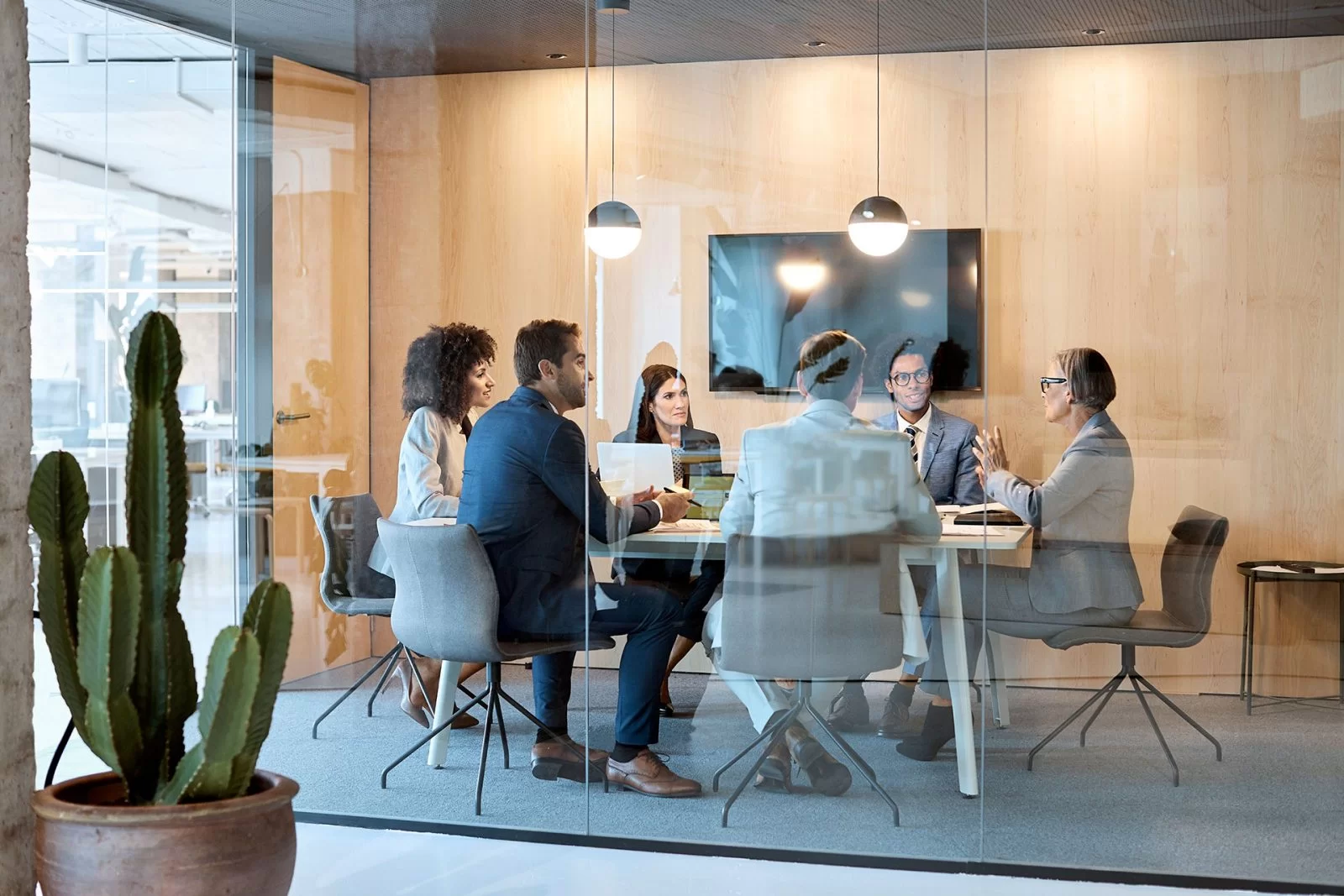 Seven people sit around a conference table in a modern glass-walled office, engaged in a business meeting. A large screen is mounted on a wooden wall behind them, and a cactus is visible in the foreground.