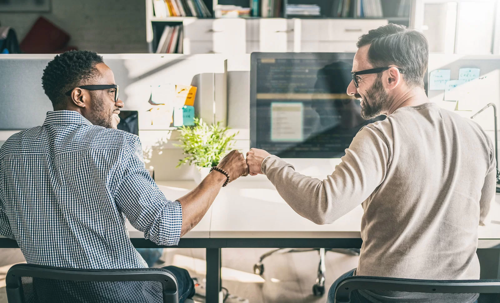 Two men sitting at desks in an office, smiling as they give each other a fist bump, with computer monitors and office supplies visible in the background.