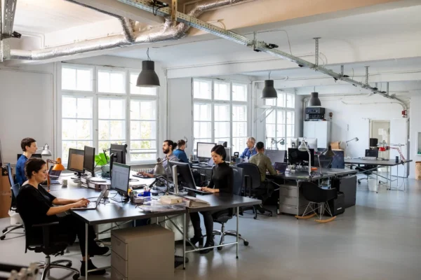 Modern open-plan office with several people working at desks on computers, surrounded by large windows, exposed pipes, and bright lighting. Some individuals are focused on screens while others interact.