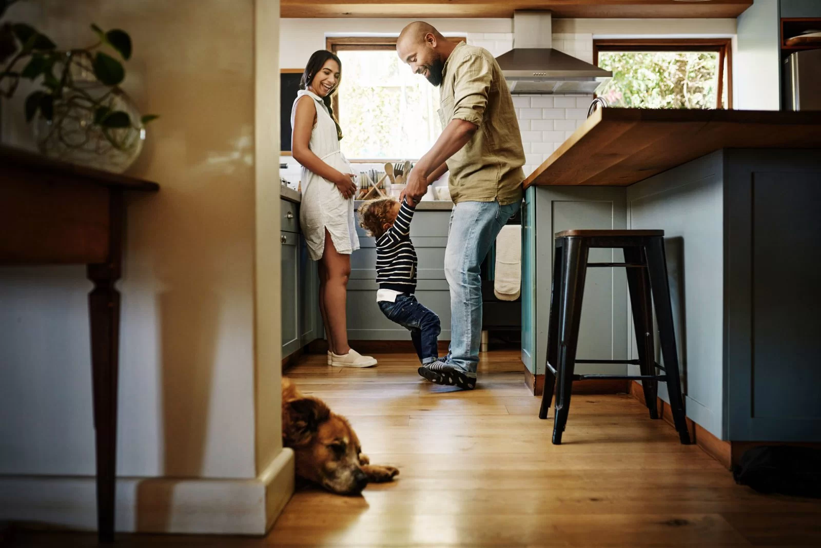 A smiling couple in a kitchen holds hands with a young child, lifting them playfully. Sunlight fills the cozy room, and a brown dog lies relaxed on the wooden floor in the foreground.