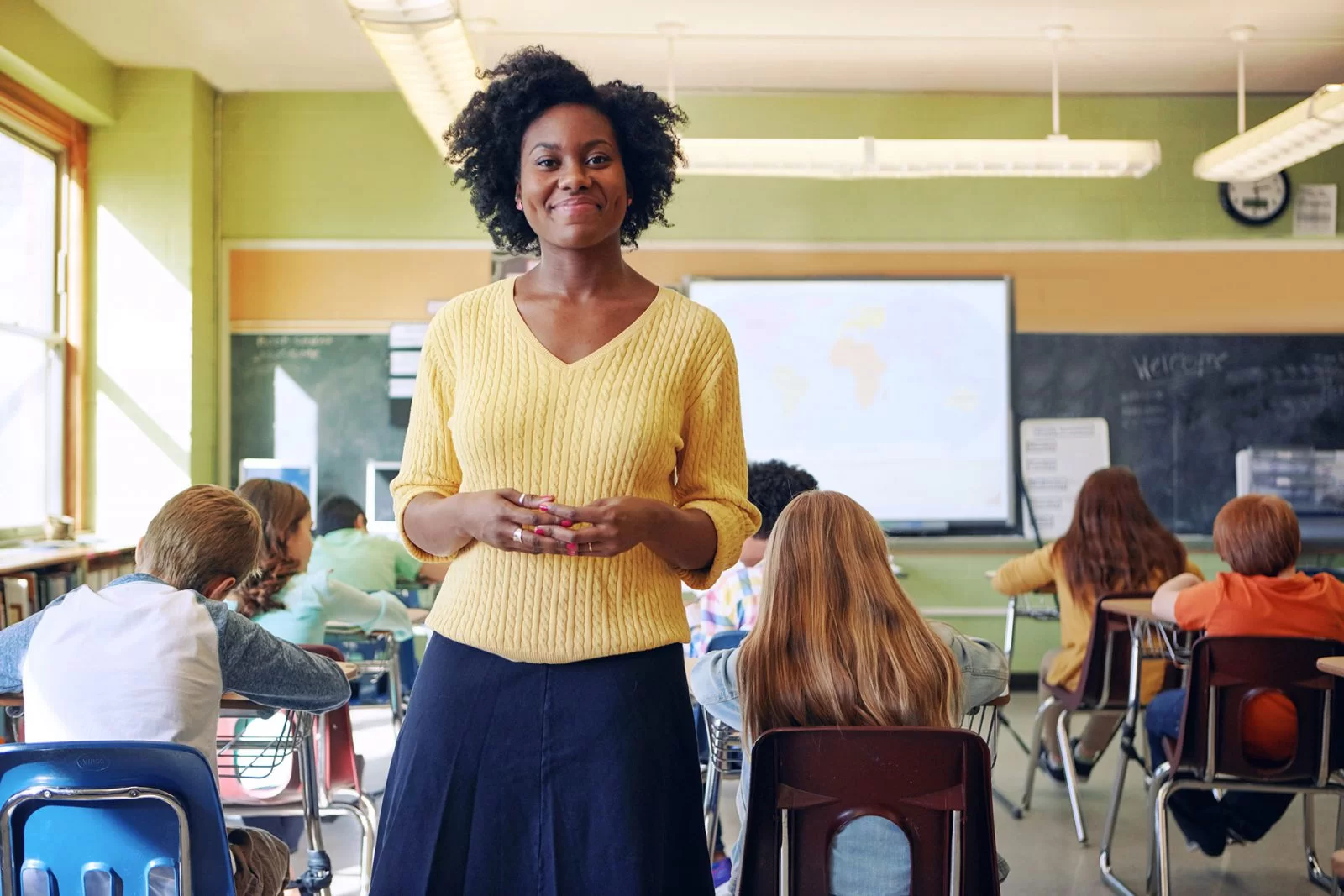 A teacher in a yellow sweater stands smiling at the front of a classroom, with students seated at desks working. The classroom has large windows, a chalkboard, and a world map in the background.