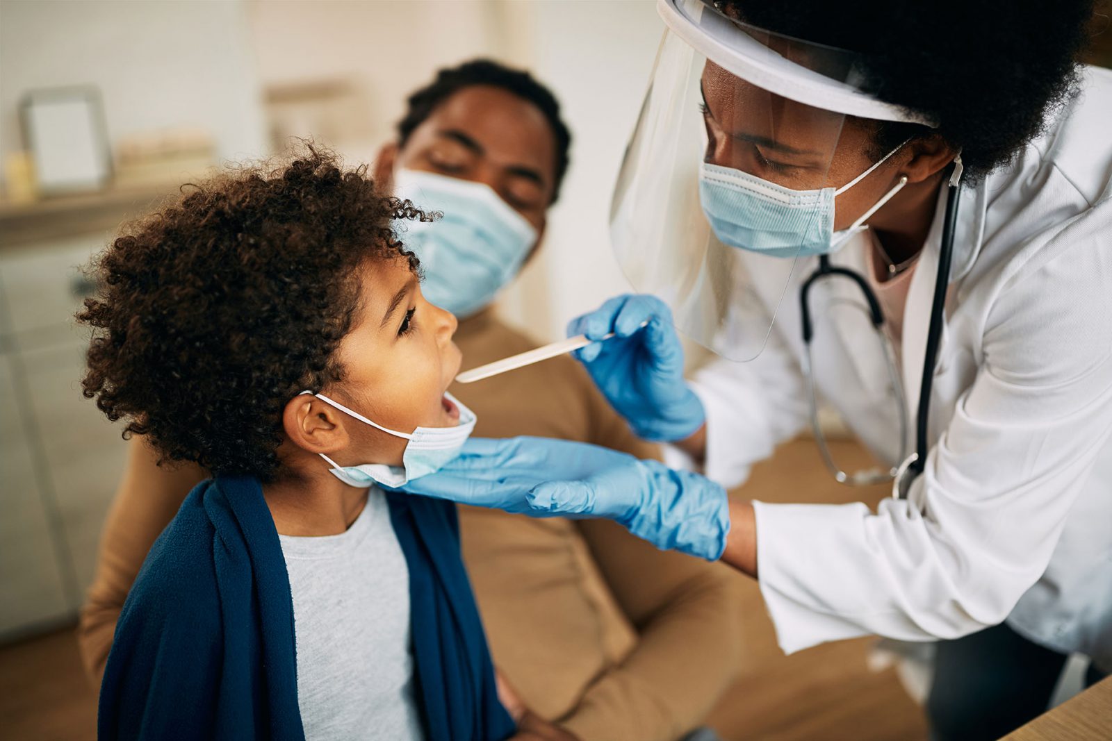 A healthcare professional in a face shield and mask examines a young boy’s throat with a swab while his guardian, also masked, looks on in a medical setting.