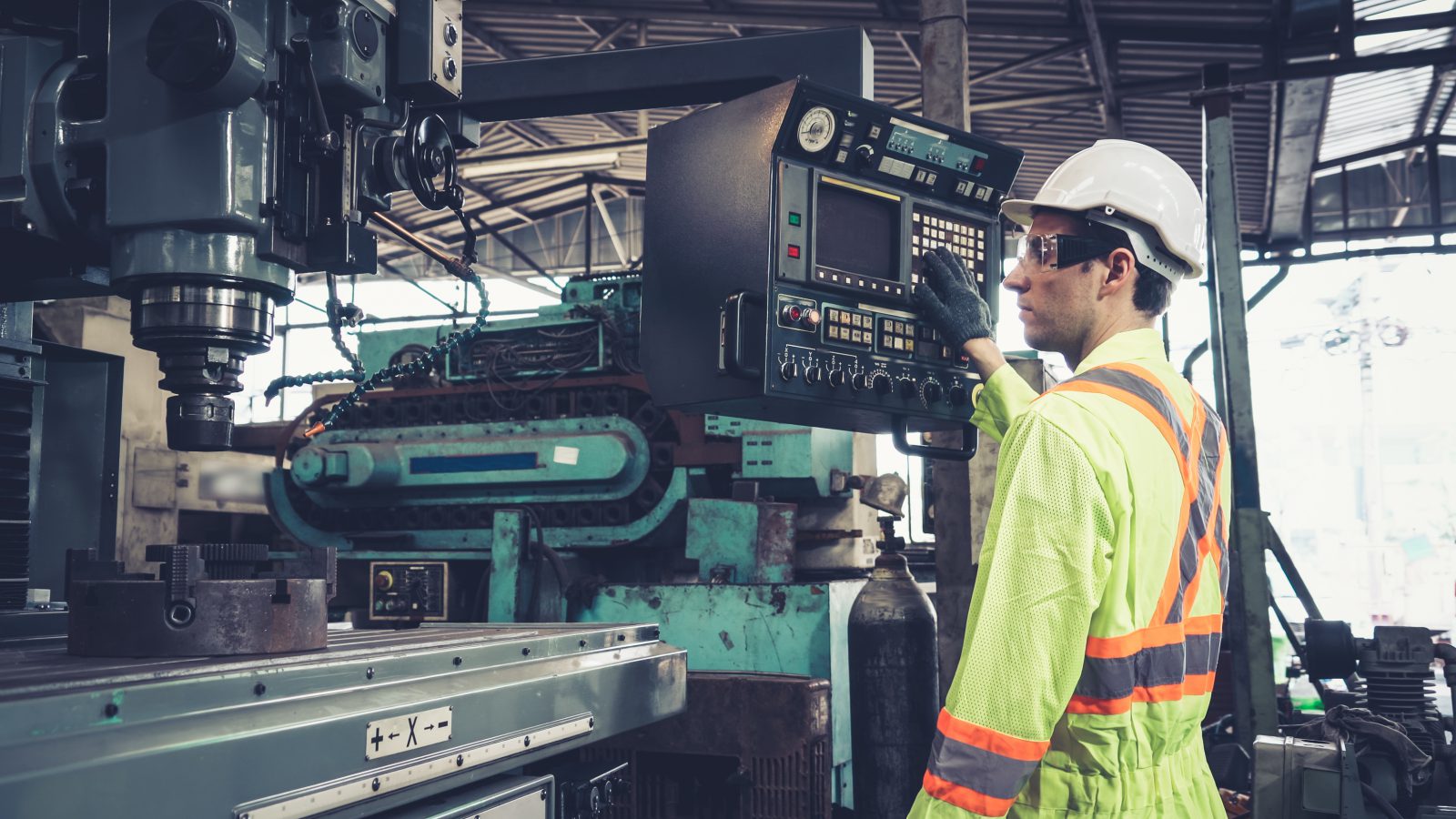 A factory worker in a hard hat and safety vest operates a control panel on a large industrial machine in a manufacturing facility.