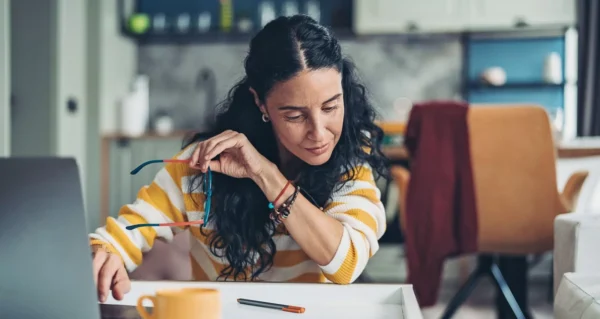 A woman with long dark hair wearing a yellow and white striped sweater sits at a desk, holding her glasses in one hand while looking at papers. A laptop, pen, and mug are on the desk in a cozy home setting.