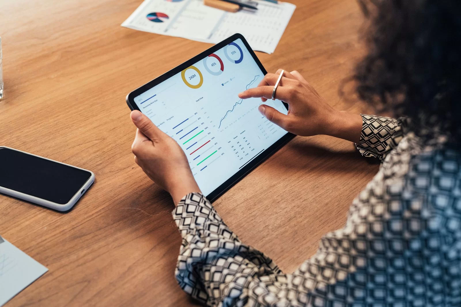 Person using a stylus to analyze graphs and charts on a tablet at a wooden desk, with a smartphone and papers nearby. The screen displays data visualizations and performance metrics.