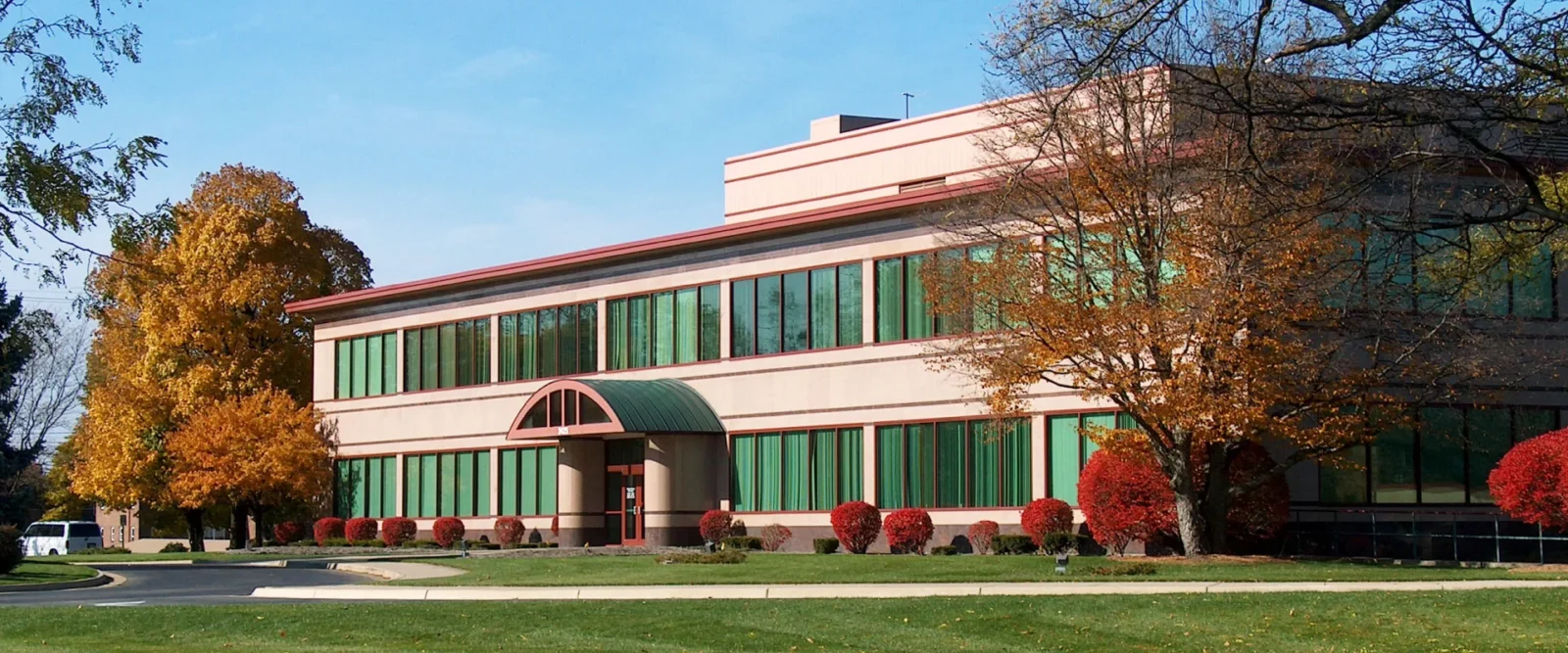 A two-story office building with large green-tinted windows, beige walls, and a green awning over the entrance. Autumn trees with colorful leaves surround the building, and a manicured lawn is in front.