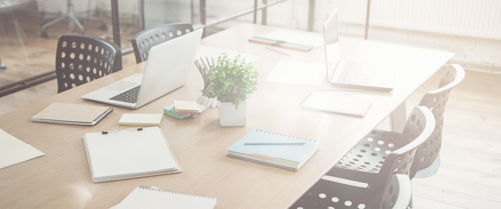 A bright, modern office meeting room with a wooden table, laptops, notebooks, clipboards, folders, a plant, sticky notes, and black chairs, all illuminated by sunlight through large windows.