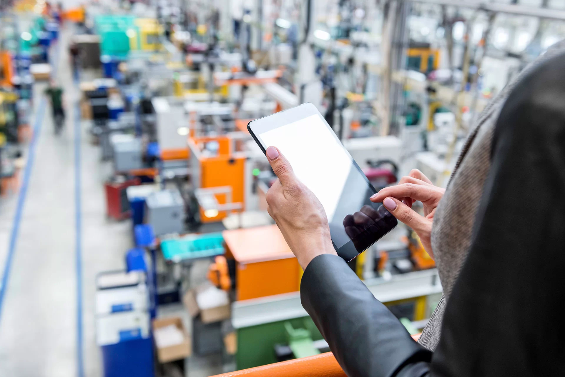 A person holding and using a tablet overlooks a busy factory floor filled with machinery, equipment, and workers.