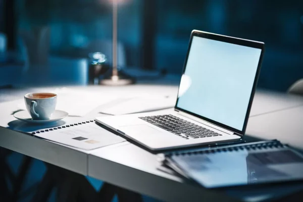 A laptop sits open on a desk next to a cup of coffee, documents, and a spiral notebook in a softly lit office setting at night.