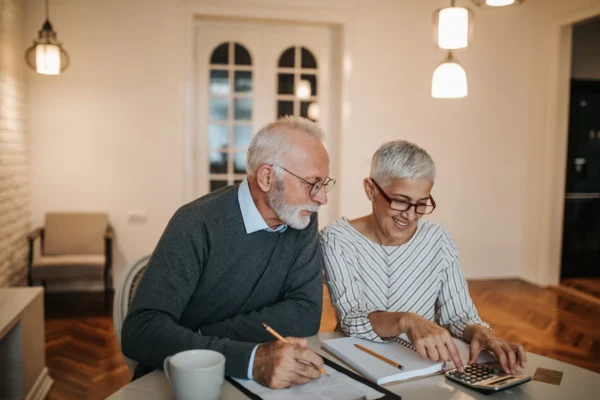 An older man and woman sit together at a table, smiling while working on paperwork. The woman uses a calculator, and the man holds a pencil. They appear to be discussing finances in a cozy, well-lit room.