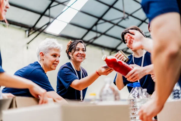 A group of people wearing blue shirts work together, smiling as they sort and pass food items like bottles and cans in a large, well-lit warehouse.