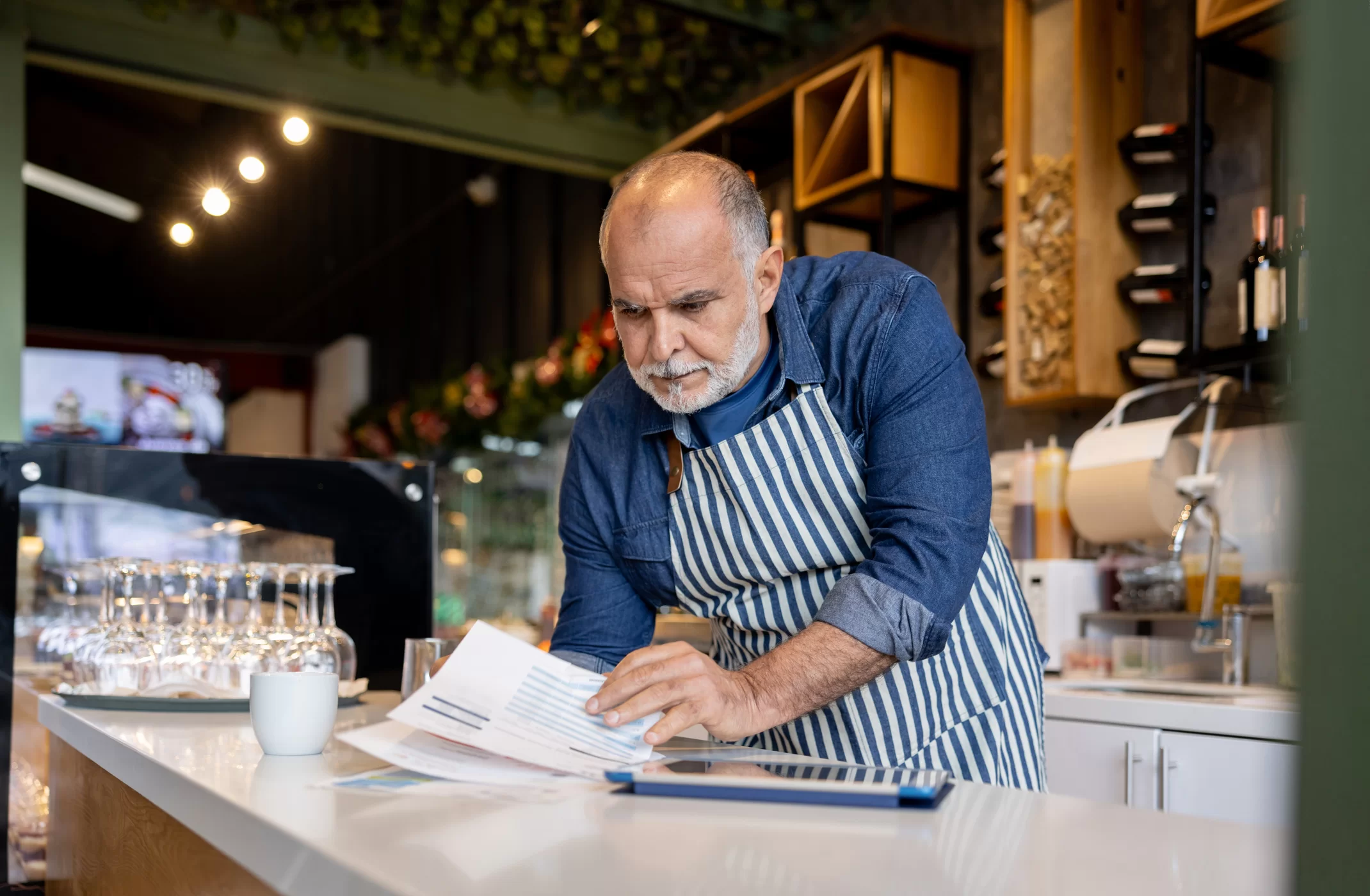 Middle-aged man wearing a striped apron reviews papers at a counter in a café or restaurant, with a coffee cup and glassware nearby. He appears focused on the documents in a well-lit, modern setting.