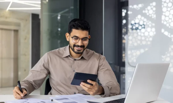A man wearing glasses sits at a desk with papers, a laptop, and a pen, smiling while looking at a tablet in his hand.