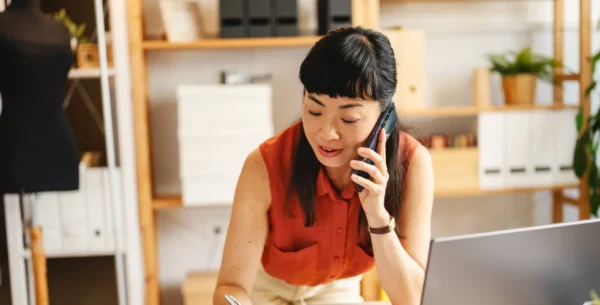 A woman in an orange sleeveless blouse talks on the phone while writing notes at a desk in a modern, well-organized office space. A laptop and shelves with boxes and plants are visible in the background.
