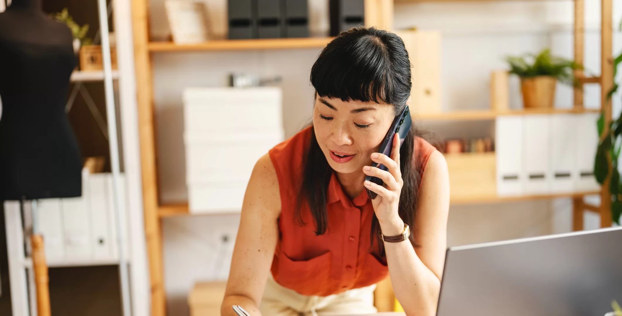 A woman in an orange sleeveless blouse talks on the phone while writing notes at a desk in a modern, well-organized office space. A laptop and shelves with boxes and plants are visible in the background.