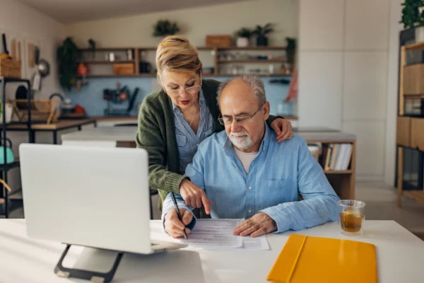 An older couple reviews documents together at a desk in a modern kitchen. The man is signing papers while the woman stands beside him, pointing at the document. A laptop and folders are on the desk.