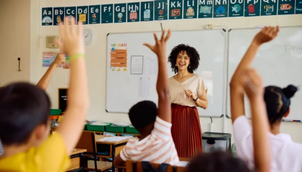 A smiling teacher stands at the front of a classroom near a whiteboard, while several students with raised hands sit at their desks. The classroom wall displays an alphabet chart above the board.