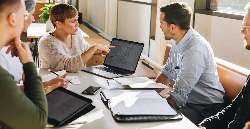 Four people sit around a table in a bright office, discussing something on a laptop. One woman points at the laptop screen while others listen attentively. Papers, a tablet, and a notebook are on the table.