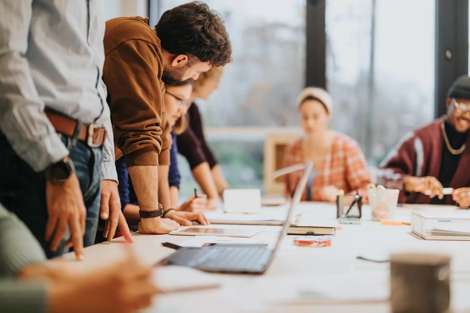 A group of people sits and stands around a table in a bright office, collaborating and discussing documents and laptops during a meeting.