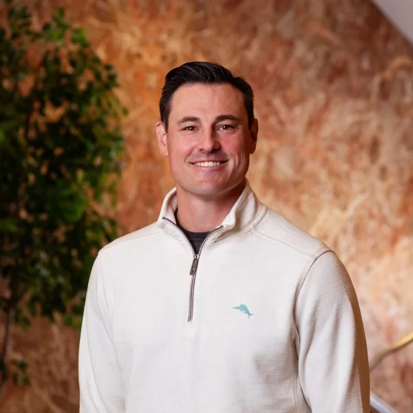 A man with short dark hair wearing a white quarter-zip sweater stands indoors in front of a textured wall with greenery to the side, smiling at the camera.