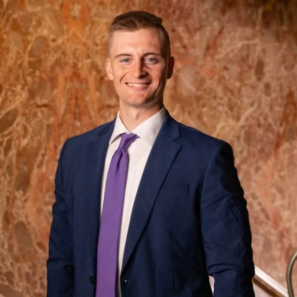 A man in a navy blue suit with a white shirt and purple tie smiles, standing indoors against a marbled orange and brown background.