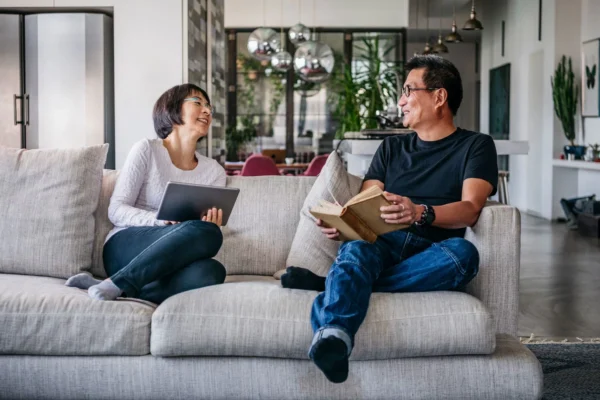Two people sit on a sofa in a modern living room, smiling and talking. One person holds a tablet, and the other holds an open book. The space is bright with large windows and contemporary decor.