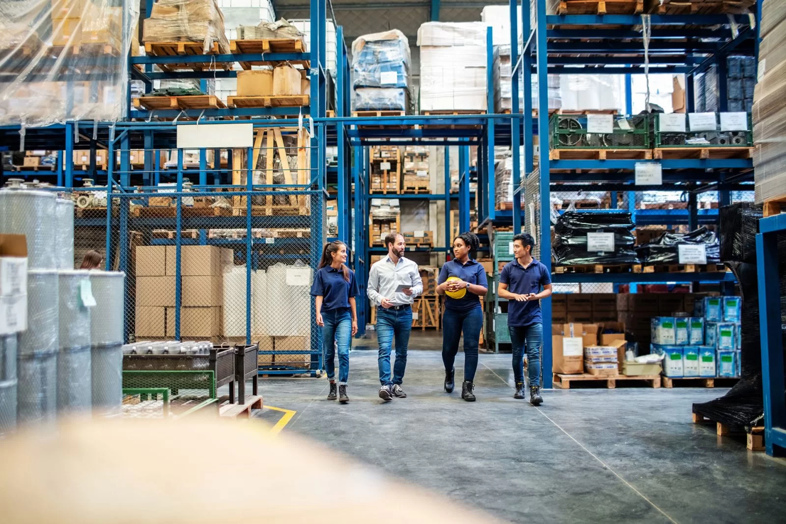 Four people in work attire walk and talk inside a large, organized warehouse filled with shelves stacked with boxes and various items. The environment is brightly lit and spacious.