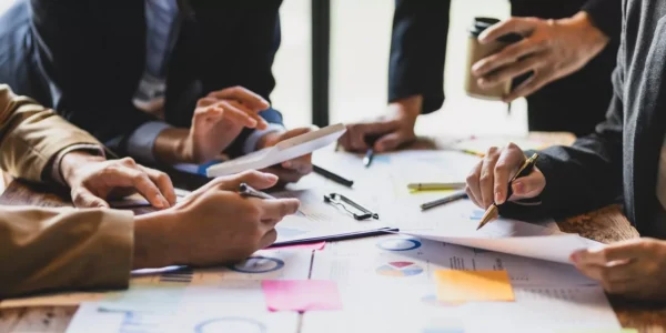 A group of people sit around a table covered with documents, charts, graphs, and sticky notes, discussing and pointing at papers and a tablet, suggesting a collaborative business meeting.