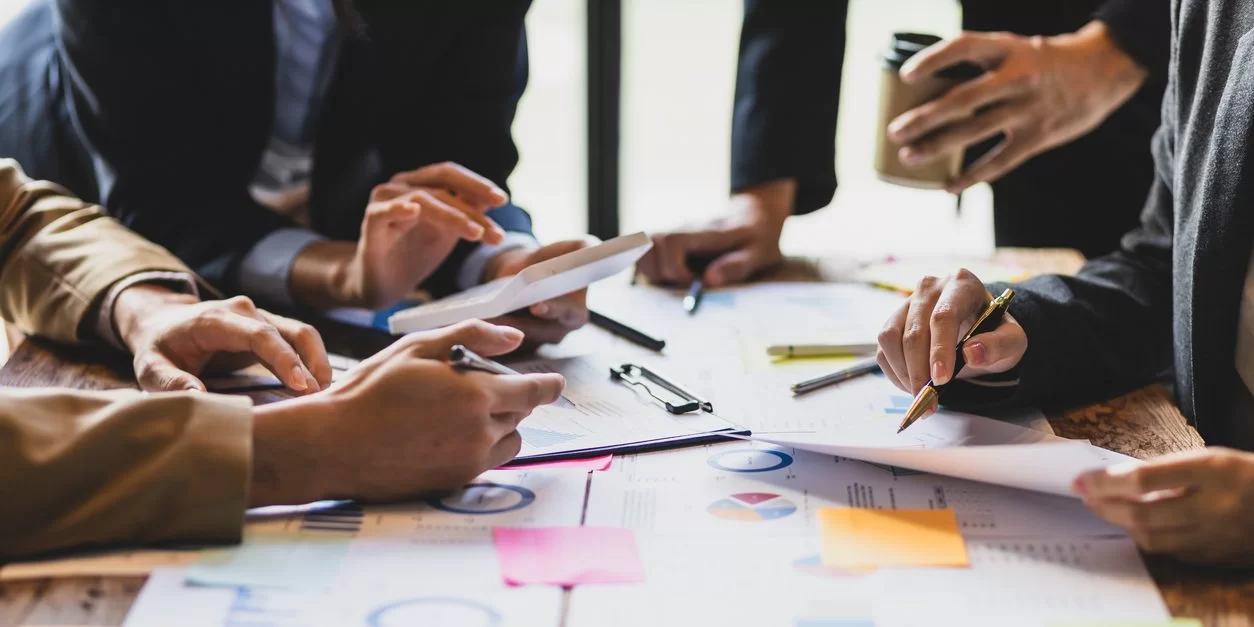A group of people sit around a table covered with documents, charts, graphs, and sticky notes, discussing and pointing at papers and a tablet, suggesting a collaborative business meeting.