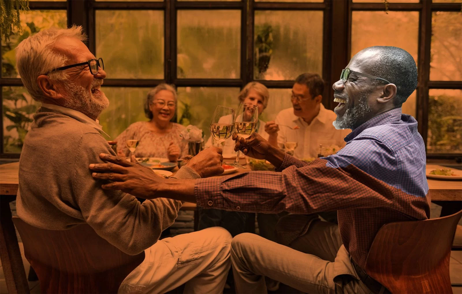 Two older men sitting at a dining table laugh and clink wine glasses, while three other older adults in the background also share smiles and enjoy a meal in a warm, cozy setting.