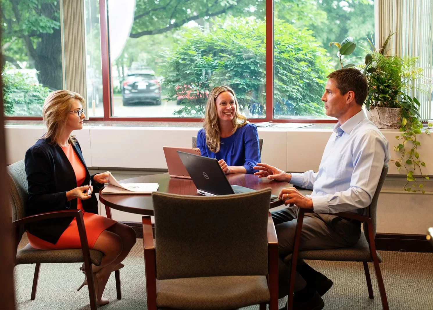 Three people sit around a small round table in an office, having a meeting about the company's privacy policy. Two women and one man are discussing details, with laptops and papers on the table. Large windows show greenery outside.