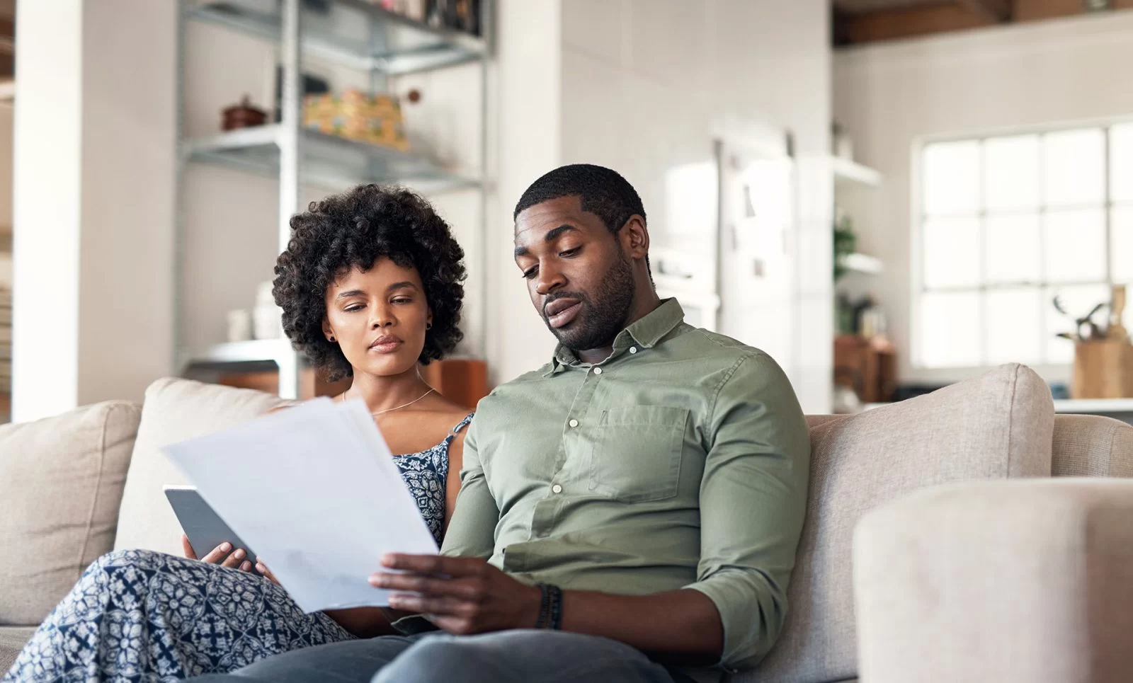A couple sits together on a couch in a bright living room, reviewing privacy policy documents and a tablet with serious expressions. Shelves and a large window are visible in the background.