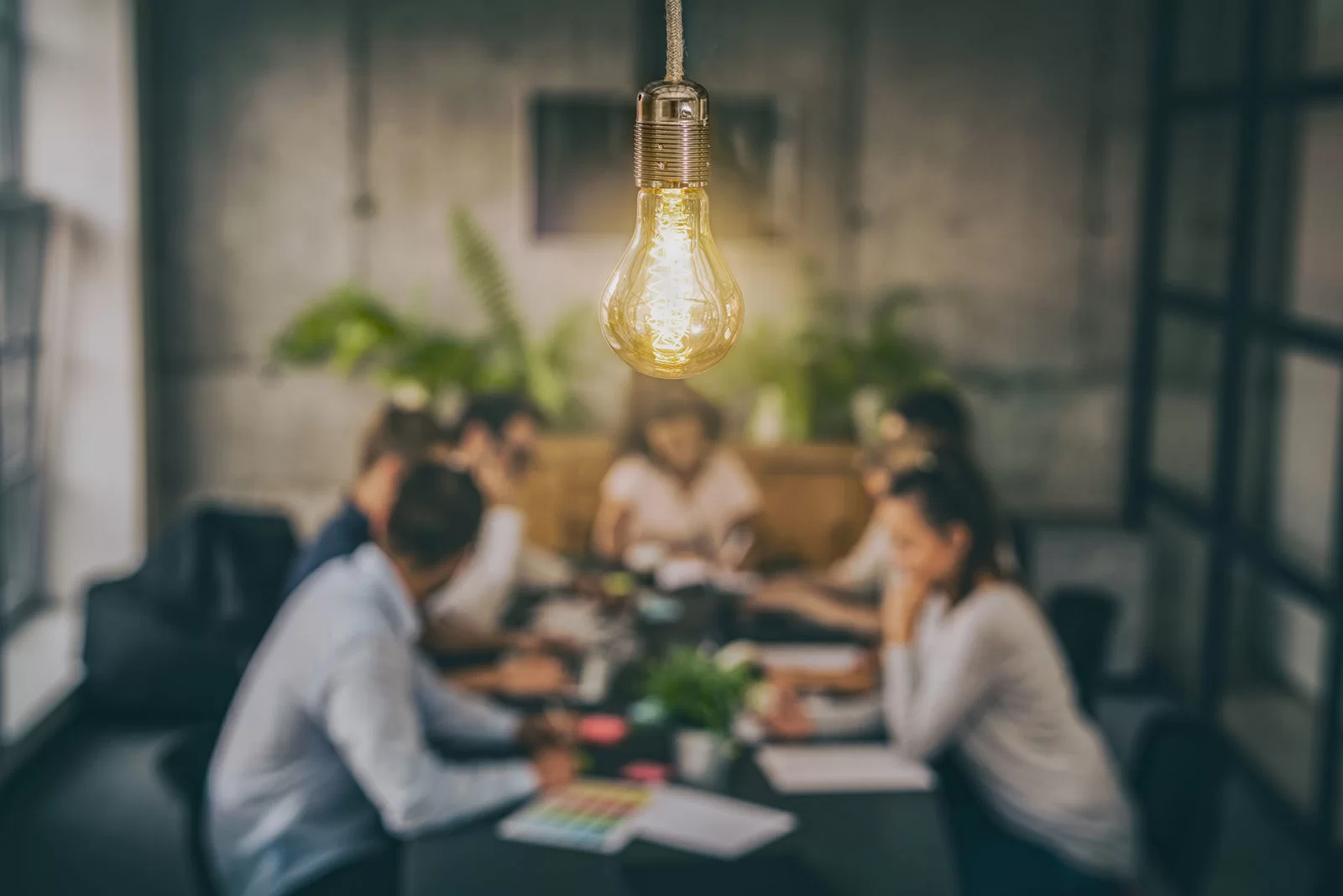 A glowing lightbulb hangs in focus in the foreground, while a group of people sit and collaborate around a table in a blurred, modern office setting with plants in the background.
