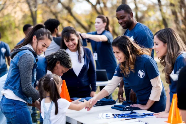 A group of volunteers at an outdoor event greet a young girl and her mother at a registration table. Everyone is smiling and interacting under trees, with shirts and lanyards displayed on the table.