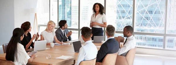A woman stands and speaks to a group of colleagues seated around a conference table with laptops, in a bright modern office with large windows overlooking city buildings.