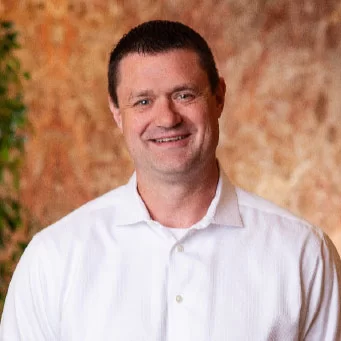 A man with short dark hair, wearing a white button-up shirt, smiles at the camera. The background is softly blurred with warm brown and orange tones, and some green leaves are visible on the left.