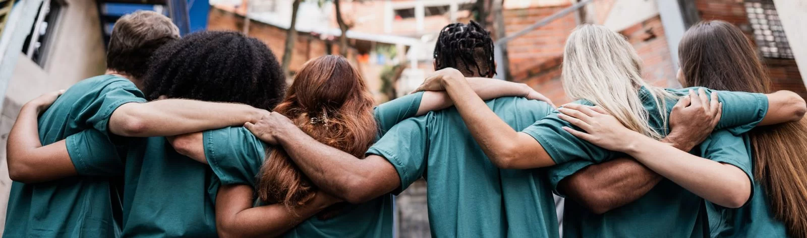 A group of six people in green shirts stand side by side outdoors with their arms around each others shoulders, facing away from the camera, showing unity and friendship.